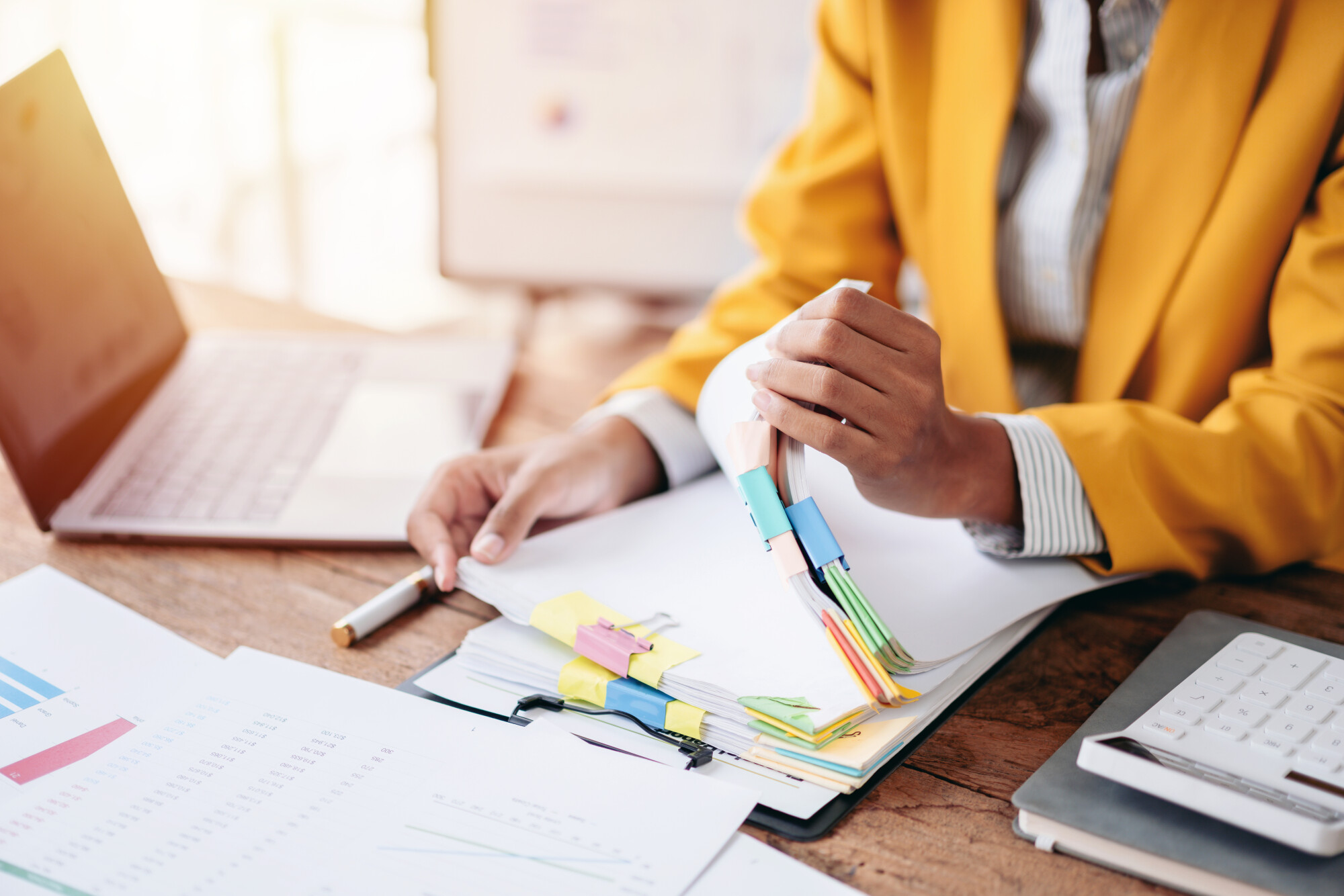 Businesswoman with yellow sports coat sorts through and analyzes financial data reports with laptop, calculator and her hands sorting documents for tax and investment planning at office