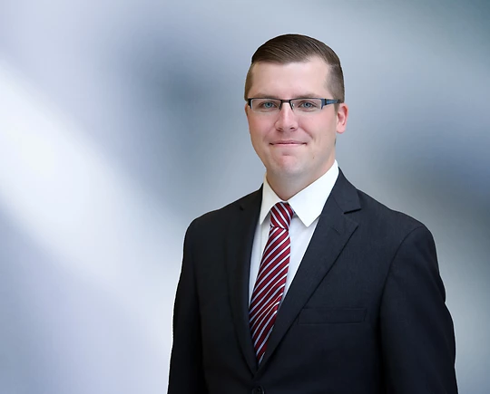 man, Taylor Gast, in a dark suit smiling at the camera, standing against a blurred blue and white background.