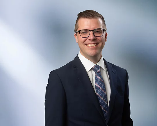 A man, Taylor Gast, in a dark suit smiling at the camera, standing against a blurred blue and white background.