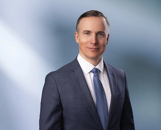 A man, Joel Farrar, in a blue suit, white shirt, and blue tie is looking at the camera, standing against a blurred blue and white background.